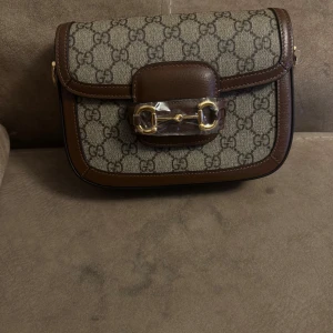 Gucci GG Supreme axelväska brun - Snygg axelväska från Gucci i klassiskt GG Supreme-mönster. Väskan är beige med bruna skinn-detaljer och guldfärgat spänne framtill. Två axelremmar medföljer, en i grönt/rött tyg och en i brunt skinn. Perfekt för dig som gillar lyxiga accessoarer.