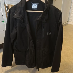 Svart vindjacka från McKinley storlek 158-164 - Svart vindjacka från McKinley med praktiska fickor och dragkedja framtill. Jackan har en stilren design med hög krage och långa ärmar, perfekt för utomhusaktiviteter. Materialet är slitstarkt syntet och jackan har diskret logga på bröstet.