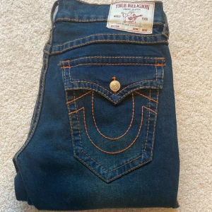 True Religion jeans med kontrastsömmar - Mörkblå jeans från True Religion med ikoniska kontrastsömmar i orange och stora bakfickor med lock och metallknappar. Klassisk femficksmodell i jeansmaterial, rak passform och låg midja. Perfekta för dig som vill sticka ut med detaljer. Säljer pga för stora
