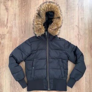 Svart pufferjacka med fuskpäls från Everest / Stadium - Svart pufferjacka från Stadium med huva och fuskpäls i bruna toner. Jackan har dragkedja framtill, ribbade muddar och två fickor med knappstängning. Yttermaterial i polyester och foder i nylon. Perfekt för kalla dagar.