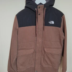 Brun och svart fleece hoodiejacka The North Face - Snygg hoodiejacka från The North Face i brun och svart med dragkedja och tryckknappar. Jackan har två stora fickor framtill, huva med snörning och mjukt fleecefoder på insidan. Perfekt för kyliga dagar och streetwear-stil. I mycket bra skick