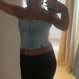 Ljusblå tubtopp med dragkedja - Säljer en ljusblå tubtopp i denim-look med råa kanter och dragkedja framtill. Toppen är axelbandslös och har en tight passform som framhäver midjan. Perfekt för dig som vill ha en edgy och trendig stil.