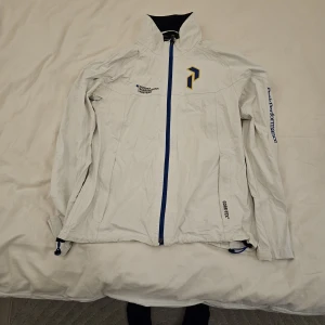 Vit golf, vind och regn jacka Peak Performance Gore-Tex bro hof slott - Vit vindjacka från Peak Performance med blå dragkedja och detaljer. Jackan är tillverkad i polyamid med Gore-Tex-membran, har hög krage och logga på bröstet samt ärmen. Broh hof nordea Scandinavian masters