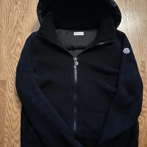 Moncler Maglione Tricot Cardigan – Svart - Säljer en Moncler Maglione Tricot Cardigan, en exklusiv hybrid mellan dunjacka och stickad tröja. Perfekt balans mellan värme, stil och komfort – idealisk för vår, höst och mild vinter.