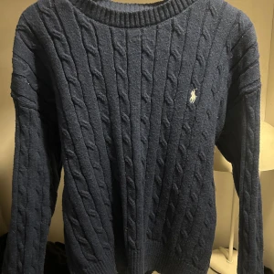 Mörkblå kabelstickad tröja Polo Ralph Lauren - Fet Kabelstickad mörkblå tröja från Polo Ralph Lauren med klassisk rund halsringning och broderad logga på bröstet. Tröjan har långa ärmar och ribbade muddar, perfekt för lager-på-lager under kyliga dagar. Storlek M men skulle säga att den passar S också.