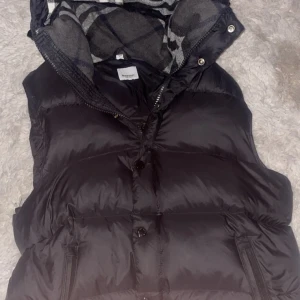 Svart dunväst från Burberry - Svart dunväst från Burberry med klassiskt rutigt foder i grått och vitt. Västen har dragkedja och knappar framtill, samt flera fickor med dragkedja. Märkets logga syns på avtagbara ärmar. Perfekt för lager-på-lager-stil under kyliga dagar.