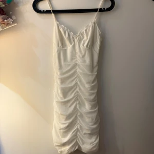 Vit kort omlottklänning - Supersöt vit kort klänning med tunna axelband och snygg rynkad effekt längs hela klänningen. Omlott-detalj upptill. Sitter tight och har en fräsch sommarkänsla, perfekt för dig som gillar en feminin och trendig stil.