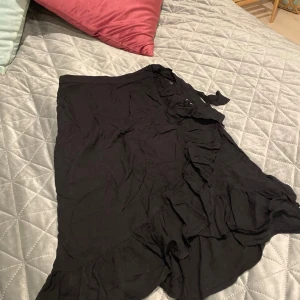 Svart omlottkjol med volang från Lindex - Svart kort omlottkjol från Lindex med snygga volangdetaljer längs kanten och knyt i midjan. Kjolen är i mjuk bomull och har en lätt och luftig känsla, perfekt för en trendig och avslappnad look.