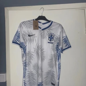 Vit Brasilien fotbollströja Nike - Snygg vit Brasilien fotbollströja från Nike med blå detaljer och grå palmbladsmönster. Tröjan har korta ärmar, broderat CBF-emblem och klassisk v-ringad krage. Perfekt för dig som vill sticka ut på planen eller på läktaren.