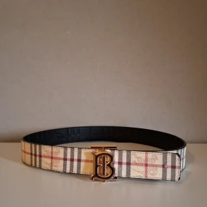 Burberry randigt skärp med guldspänne 2 sidig. - Säljer ett Burberry bälte med klassiskt rutigt mönster i beige, svart och rött på ena sidan och svart präglad skinn på andra. Snyggt guldigt TB-spänne framtill. Perfekt accessoar för att lyfta din outfit med lyxig känsla och ikonisk design. Vill man ställa frågor och/eller diskutera om priset så är det bara att kontakta mig.