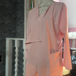 Ljusrosa omlottklänning med knytband - Säljer en ljusrosa omlottkappa med långa ärmar och praktiska fickor framtill. Kappan har ett snyggt knytband i midjan som ger en fin siluett och en stilren look. Perfekt för dig som gillar enkel men trendig design.