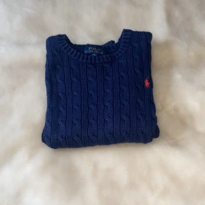 Marinblå kabelstickad tröja Polo Ralph Lauren - Kabelstickad marinblå tröja från Polo Ralph Lauren med rund hals och röd broderad logga på bröstet. Tröjan har långa ärmar och ribbade muddar vid ärmslut och nederkant. Perfekt för lager-på-lager och snygg till jeans.
