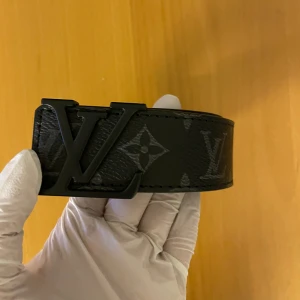 Svart skärp med monogram - Säljer ett svart bälte i skinn med ikoniskt monogrammönster och ett -spänne i mörk metall. Bältet har en klassisk och stilren look som lyfter vilken outfit som helst. Kommer med originalask och kvitto.