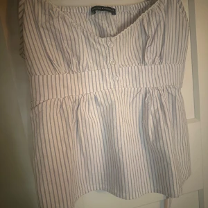 Randig blus från Brandy Melville - Supersöt blus från Brandy Melville i vit och ljusblå randigt mönster. V-ringad med tre knappar framtill, smockad midja och knytband bak. I onesize, priset kan såklart diskuteras 🩷