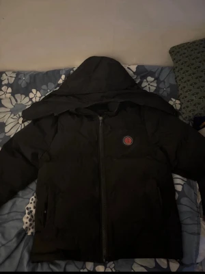 Svart pufferjacka från Trapstar - Svart pufferjacka från Trapstar med huva och dragkedja framtill. På bröstet finns ett runt Trapstar-märke och på ryggen står det 'TRAPSTAR' i röd broderad text. Jackan har en loose passform och är perfekt för kalla dagar.