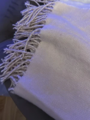 Beige halsduk med fransar - Säljer en stilren beige väst i mjuk ull med fransar längs kanterna. Västen har en enkel och klassisk design utan mönster eller knappar, perfekt för att lägga till en extra touch till din outfit.