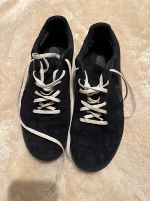 Svarta futsal skor - Svarta futsal skor från Nike med vit snörning och rund tå. Skorna har en kombination av mocka och mesh på ovansidan samt en svart Nike-logga på sidan. Yttersulan är svart med mönstrad struktur för extra grepp. 