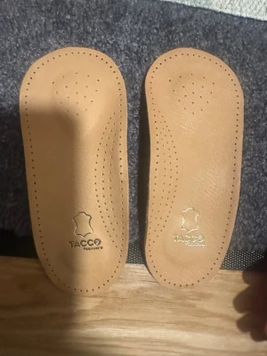 Beiga skoinlägg  hålfotsstöd i skinn från Tacco - Säljer ett par beige skoinlägg från Tacco, tillverkade i mjukt skinn med perforerade hål för extra ventilation. Perfekta för att ge extra komfort i dina skor. Undersidan har klisteryta för att hålla inläggen på plats.aldrig använda. Passar stl 40-42 ( stl M). 