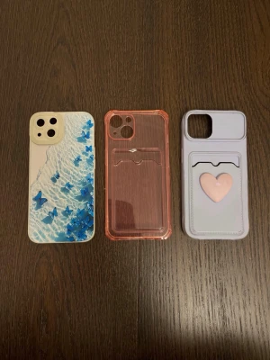 Apple mobilskal set med olika mönster - Apple mobilskal i set om tre stycken med olika mönster och färger. Ett skal har blå fjärilar på vit bakgrund, ett är genomskinligt rosa med kortfack, och ett är lila med ett rosa hjärta och kortfack. Skalen är avsedda för mobiltelefon och erbjuder skydd samt möjlighet att förvara kort. iPhone 13.