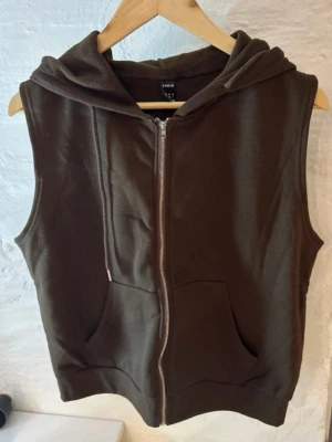 Brun hoodie-väst från SHEIN - Brun väst med huva från SHEIN, tillverkad i mjuk bomull. Västen har dragkedja framtill, två fickor och en avslappnad passform. Perfekt att slänga över en t-shirt för en chill look. Snygg och enkel design utan mönster.