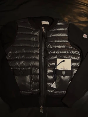 Moncler Cardigan  - Säljer en svart pufferjacka från Moncler med glansig framsida och stickade ärmar. Jackan har två fickor med knappstängning, dragkedja framtill och Moncler-logga på ärmen. Perfekt för dig som vill ha en stilren och modern look.