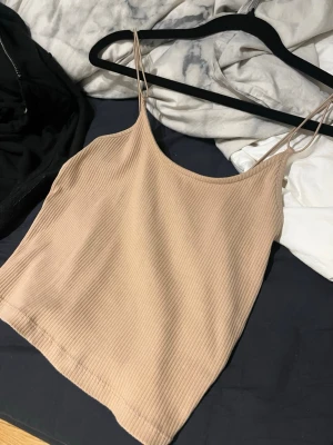 Beige ribbad linnetopp med smala band - Säljer en beige ribbad linnetopp med smala axelband. Toppen har en enkel och stilren design med rundad halsringning och ärmlös passform. Perfekt för varma dagar eller som layering-piece. Materialet är mjukt och stretchigt för skön komfort. Storlek S/M