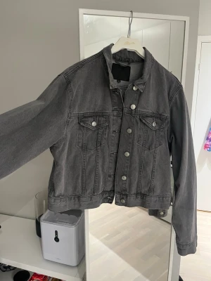 Mörkgrå jeansjacka med knappar - Säljer en mörkgrå jeansjacka med klassisk krage och silverfärgade knappar. Jackan har två bröstfickor med lock och knapp, samt raka sömmar som ger en snygg och stilren look. Perfekt att slänga på över en hoodie eller t-shirt.