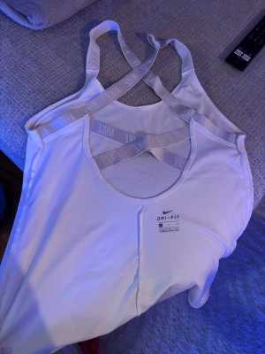 Vit Nike Dri-Fit linnetopp - Säljer en vit sportig linnetopp från Nike med Dri-Fit material. Toppen har tunna axelband med Nike-logga och snyggt korsad rygg. Perfekt för träning och riktigt skön passform.