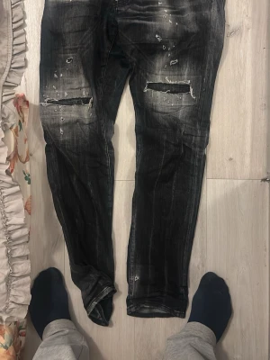 Svarta Dsquared2 Cool Guy jeans, stl 50 - Säljer ett par svarta Dsquared2 Cool Guy jeans i storlek 50. Jeansen har slitna detaljer, trasiga partier och en tvättad look som ger en rå och edgy vibe. Modellen är smalare i benen och har klassiska fem fickor. Tillverkade i Italien av bomull.