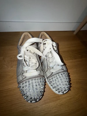 Christian Louboutin - Säljer ett par silverfärgade sneakers från Christian Louboutin med glittrigt tyg och vita snören. Insidan är i beige skinn och yttermaterialet har en glittrig finish.