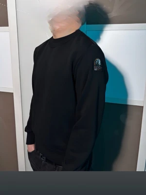 Svart sweatshirt från Parajumpers - Svart sweatshirt från Parajumpers med rund hals och ribbade muddar. Snyggt broderat PJS-märke på vänster ärm. Tröjan har en clean och stilren look, perfekt för dig som gillar streetwear och vill ha något enkelt men ändå med en exklusiv touch.