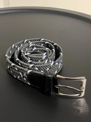 Flätat bälte 100 cm - Flätat bälte i färgen grå med blåa och vita detaljer. Det är 100 cm långt, men går att stretcha ut. Aldrig använt.