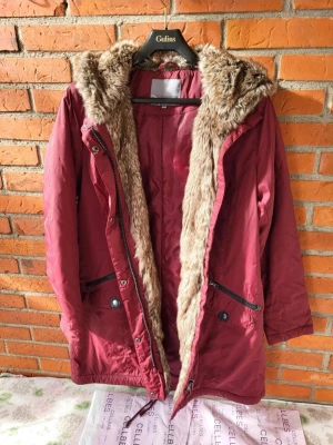 Vinröd parkajacka med fuskpäls 38-40 - Parkajacka i vinröd färg från Cellbes med mysig fuskpäls runt huvan och insidan. Jackan har dragkedja, knappar, snörning i midjan och två stora fickor med både dragkedja och knapp. Perfekt för kalla dagar och har långa ärmar.