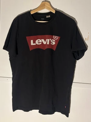 Svart Levi's t-shirt med röd logga - Säljer en svart t-shirt från Levi's i storlek L med den klassiska röda Levi's-loggan tryckt på bröstet. T-shirten har rund hals och korta ärmar, tillverkad i mjuk bomull som är skön mot huden. Perfekt för dig som gillar streetwear och ikoniska märken.