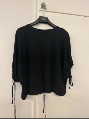 Svart topp med snörning  - Säljer en svart topp från Dr Denim i storlek XS. Toppen har loose passform och är tillverkad i mjuk bomull. Snygga snörningsdetaljer på ärmarna och nedtill ger en cool look. Perfekt att styla med jeans eller kjol för en chill vibe.