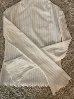 Vit ribbad polotopp med lång ärm - Säljer en vit polotopp med ribbad struktur och långärmade ärmar. Toppen har vågiga kanter vid ärmslut och nederkant som ger en cool touch. Materialet känns mjukt och har ett lätt glansigt utseende. Perfekt att styla med jeans eller kjol.