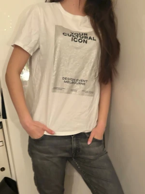 Zara t shirt - Säljer en vit t-shirt från zara som inte finns längre med svart texttryck och en stor silvrig ruta på bröstet där det står 'YOUR CULTURAL ICON' och 'DESIGN EVENT MELBOURNE'. T-shirten har rund hals och korta ärmar, och är gjord i mjuk bomull. Perfekt för dig som gillar grafiska prints och enkel stil.