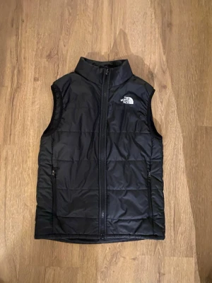 Svart dunväst från The North Face - Säljer en svart dunväst från The North Face med hög krage och dragkedja framtill. Västen har två sidofickor med dragkedja och ett stilrent, quiltat mönster. Loggan syns tydligt på bröstet och ryggen. Perfekt för lager-på-lager under kyliga dagar. Storleken är 160