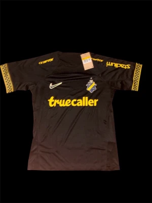 AIK tröja - Bilderna visar en svart AIK fotbollströja med gula detaljer och loggor från Nike, Truecaller och SVEA. Ingen sko finns med på bilderna.
