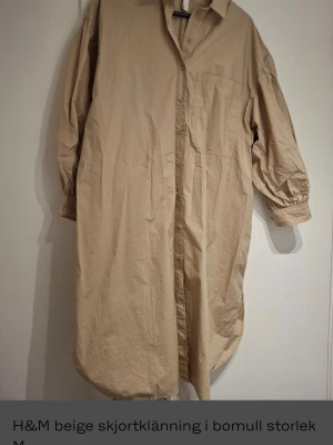 Beige skjortklänning från H&M - Beige skjortklänning från H&M i storlek M, tillverkad i 100% bomull. Klänningen har lång ärm med knappslut, klassisk skjortkrage och knappar framtill. Modellen är lång och har en loose passform med bröstficka och rundad nederkant.