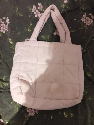 Ljuslila quiltad axelväska - Quiltad axelväska i ljuslila färg med mjuka, vadderade handtag. Väskan har ett rutmönster och är tillverkad i syntetmaterial. Perfekt storlek för att få plats med allt du behöver och har en trendig, puffig look. Den har några små fläckar på som jag visat på bilderna.