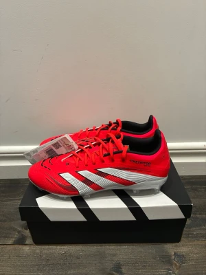 Adidas Predator Elite FG J fotbollsskor - Säljer ett par Adidas Predator Elite FG J fotbollsskor i röd och vit färg med svarta detaljer. Skorna har snörning, låg profil och mönstrad ovandel med Strikeskin-teknologi. Sulan är vit med svarta Predator-tryck och dobbar för gräsplan. Perfekt för dig som vill sticka ut på planen.