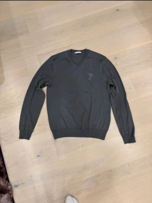 Mörkgrå V-ringad tröja Versace - Snygg mörkgrå V-ringad tröja från Versace Collection med diskret broderad Medusa-logga på bröstet. Tröjan har långa ärmar, ribbade muddar och är tillverkad i mjuk bomull. Perfekt för dig som gillar stilrena och exklusiva plagg. ÄKTA PLAGG!!! För att kolla äkthet, gå till certilogo.com och lägg in koden som visas bredvid "CLG". Tyvärr ingen kvitto eftersom fick den som present. Inga defekter finns det. 