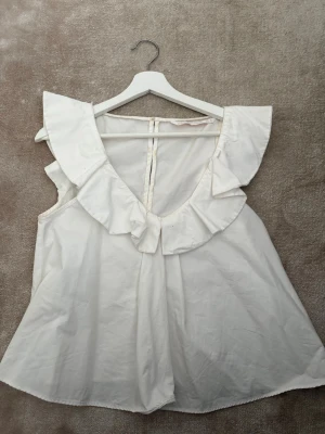 Vit blus med volang från Zara - Säljer en vit blus från Zara Trafaluc collection i storlek S. Blusen har bred volang runt halsringningen och är ärmlös med en lätt A-linjeformad siluett. .