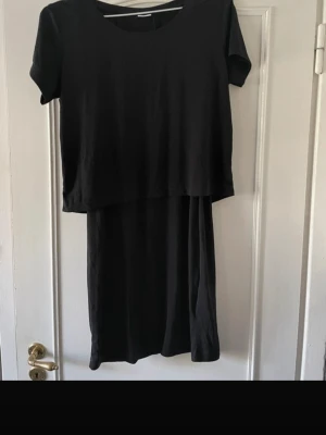 Svart kortklänning från Jacqueline de Yong - En enkel och stilren svart kortklänning från Jacqueline de Yong i storlek medium. Klänningen har rund halsringning och korta ärmar, med en rak och loose passform. Tillverkad i mjuk bomull som känns skön mot huden. Perfekt för dig som gillar minimalistisk stil.