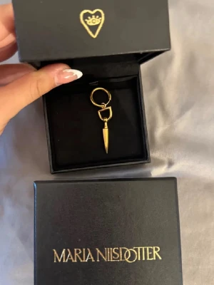 Guldörhänge från Maria Nilsdotter - Säljer ett snyggt örhänge från Maria Nilsdotter i guldfärgad metall. Designen är modern med en spetsig hängande detalj och rund ring. Perfekt för dig som gillar statement-smycken och vill ha något unikt i smyckeslådan. Skriv för fler bilder! Boxen kommer med