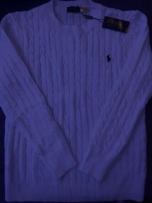 Vit kabelstickad tröja Polo Ralph Lauren - Vit kabelstickad tröja från Polo Ralph Lauren i storlek S. Tröjan har rund hals, långärmade ärmar och den klassiska broderade loggan på bröstet. Mjuk och skön stickad bomull som passar perfekt till svala dagar. Snygg ribbad kant vid hals, ärmslut och nederkant.