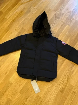 Svart pufferjacka från Canada Goose - Svart pufferjacka från Canada Goose med huva och klassisk logga på ärmen. Jackan har raka linjer, dragkedja och stora fickor framtill. Perfekt för kalla dagar och har en clean, stilren look som funkar till streetwear. Fusion fit.