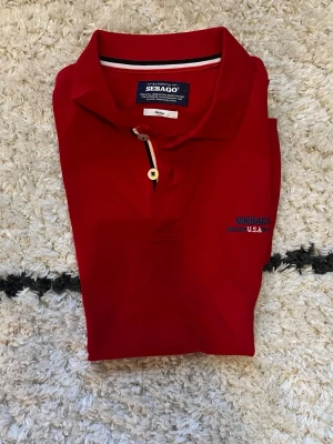 Röd pikétröja från Sebago - Klassisk röd pikétröja från Sebago med krage och knappslå med två knappar. Diskret logga broderad på bröstet och snygga kontrastdetaljer vid knapparna. Perfekt till jeans eller shorts för en avslappnad stil. Storlek small. Skriv om funderingar eller om du vill ha fler bilder.