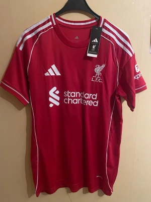 Liverpool hemmatröja – Mohamed Salah #11 - Säljer Liverpools klassiska röda hemmatröja med Mohamed Salah och nummer 11 på ryggen. Tröjan är i storlek M, med taggarna kvar. Stilren design med Standard Chartered som sponsor framtill samt Premier League-detaljer på ryggen. Tillverkad av Adidas med lätt och bekvämt material som passar både match och vardagsbruk. Ett perfekt plagg för Liverpool-supportern eller samlaren som vill ha en tröja med en av klubbens största profiler.